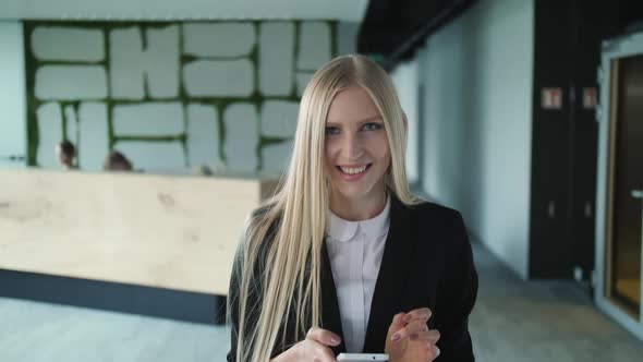 Cheerful Young Woman with Phone in Office. Pretty Blond Woman with Long Hair Wearing Elegant Black