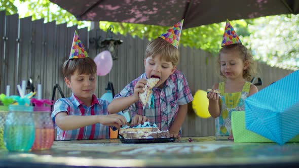 Three happy kids in party hats eating birthday cake with hands outdoors