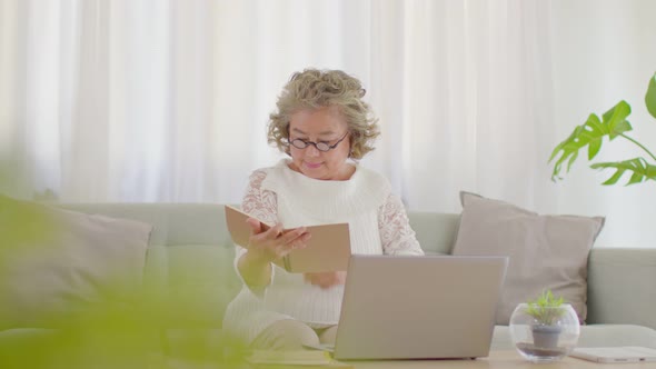 Asian senior woman hold book and learning with computer laptop sit on sofa at home