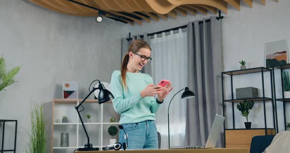 Girl in Casual Clothes Standing Near Table in Living-Room and Uses Her Mobile to Watch Funny Videos
