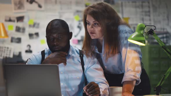 Afro Man with Holsters and Cigarette with Woman Colleague Looking at Laptop Screen in Police Office