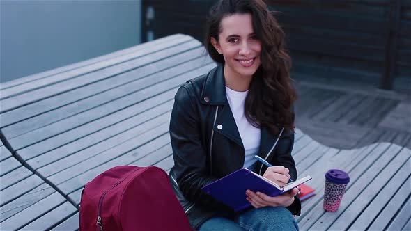 Female Student Writing in Notebook Sitting Outside on the Modern Wooden Bench