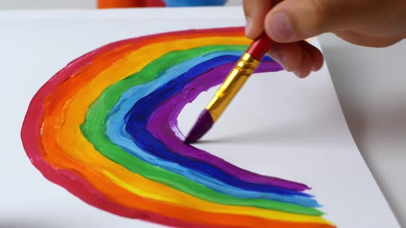 A child girl sitting at home during the quarantine period paints a multi-colored rainbow with paints