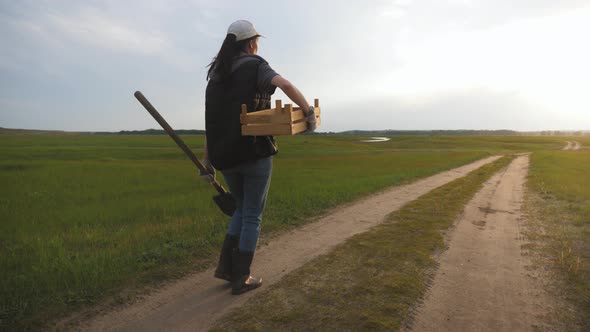 Young Gardener Woman Wearing Gumboots and Harvesting Tools in Hand Goes Along a Village Road at