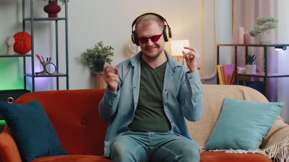 Overjoyed Young Man in Wireless Headphones Dancing Singing on Cozy Couch in Living Room at Home