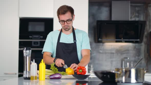 Cheerful Young Man in Apron Preparing Healthy Meal with Vegetables in Kitchen.