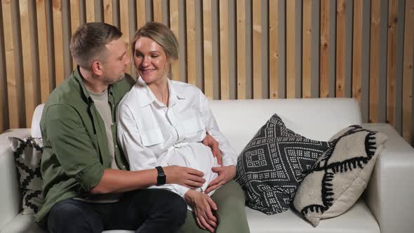 Man Strokes Pregnant Lady Tummy Sitting on White Sofa