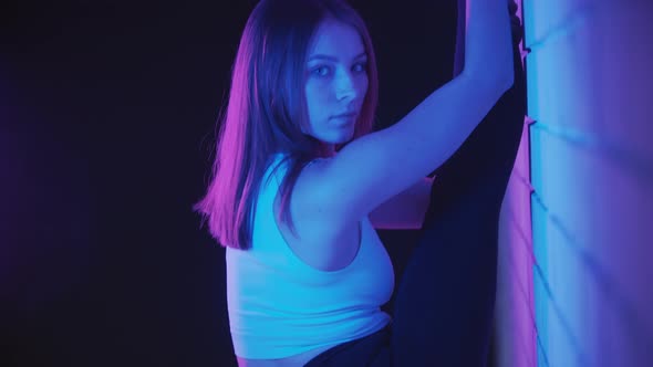 Young Woman Exercising in Neon Lighting  Stretches Her Legs Against the Wall and Looks in the Camera