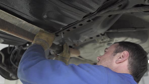 Experienced Auto Mechanic in a Blue Uniform Standing Under the Car with a Wrench and Tightening the