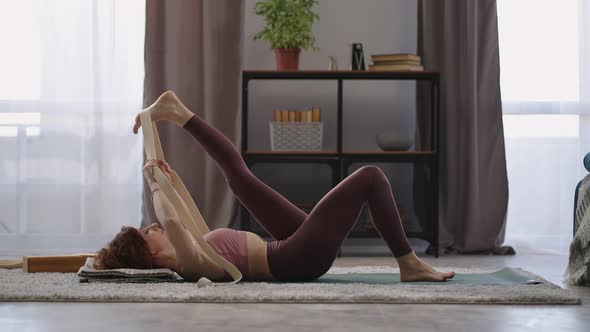 Woman is Performing Gymnastics for Flexibility Using Elastic Band for Stretching Leg Lying on Floor