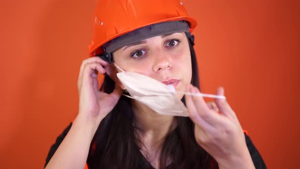 Female Construction Worker in Overalls Putting on Medical Mask on Face on Orange Background