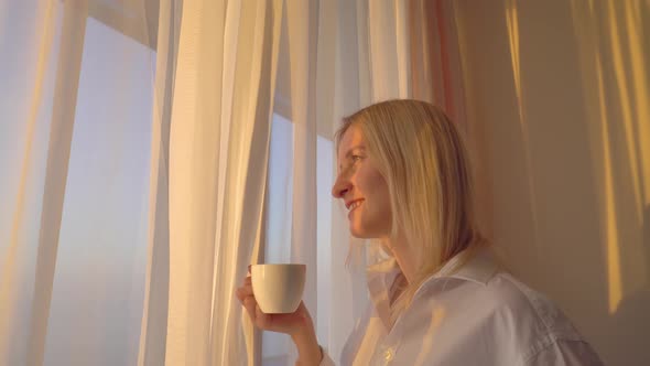 A thoughtful young blonde with a cup of coffee or tea looks out the window,