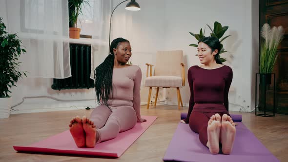 Two Diverse Women Friends Warming Up Their Feet and Talking Exercising on Fitness Mats at Home
