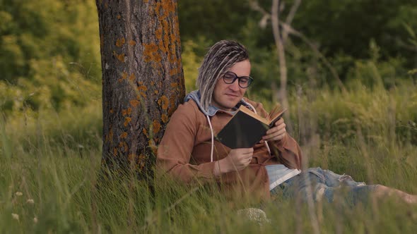 A Man in Casual Clothes and Glasses Reads Literature Under a Tree