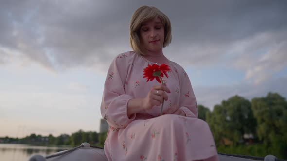 Sad Thoughtful Transgender Woman Caressing Red Flower Petals Sitting in Boat on Lake