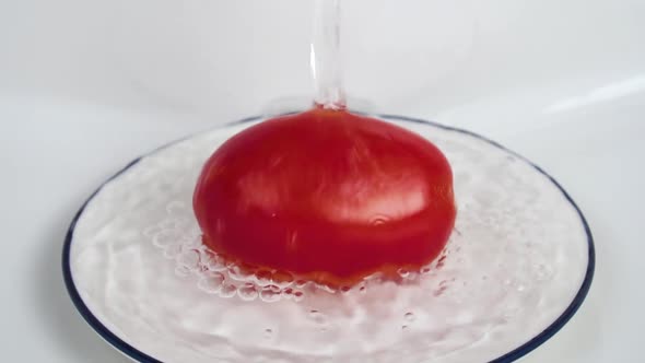 Water pours over a red ripe tomato in a white plate in the sink