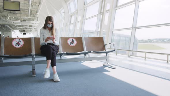 Young Woman in a Protective Face Mask Sitting in the Airport Lounge and Uses Her