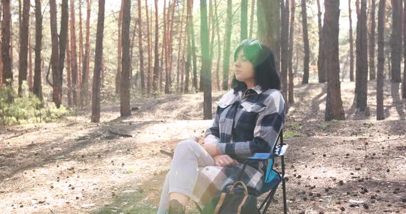 Woman deep breathing resting, sitting in chair with tourist backpack in pine forest in sun shin