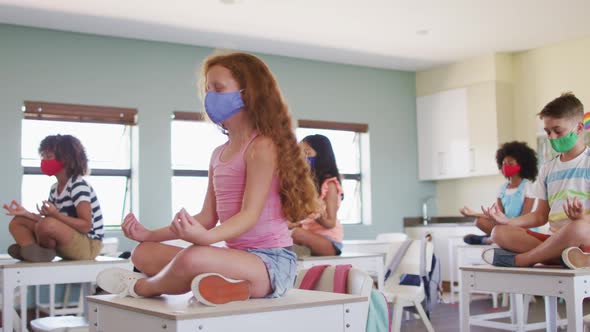 Group of kids wearing face masks practicing yoga while sitting on top of desks at school