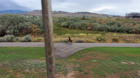 Senior man riding a bike along a paved path by a river with mountains and overcast sky - aerial foll