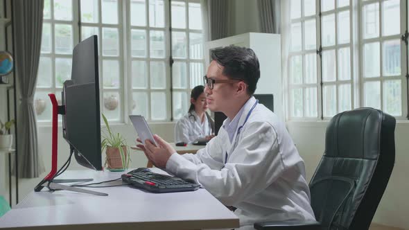 Asian Man Doctor Is Using Tablet While Work With Desktop Computer In Workplace. Medical Concept