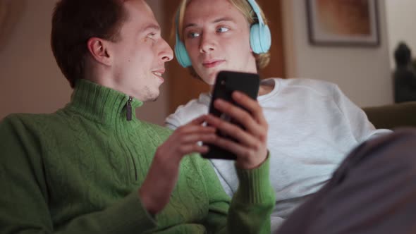 Positive guy showing something on phone to his boyfriend in headphones