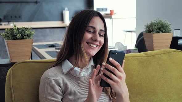 Beautiful Caucasian Woman in Beige Sweater Sitting on a Green Sofa and Using Smartphone or Cell