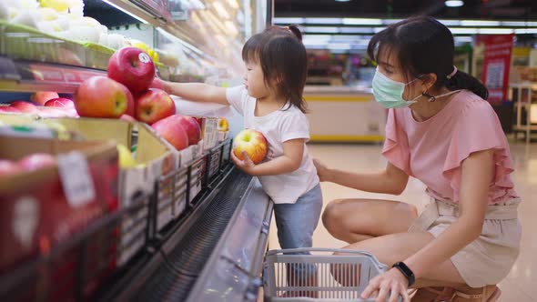 Asian young mother wear mask, hold grocery basket with her little kid child walking in supermarket.