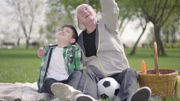 Old Man Sitting with His Grandson on the Blanket in the Park, Pointing Up