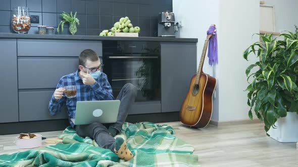 Student in protective medical face mask trying to drink tea while sitting on the floor with laptop