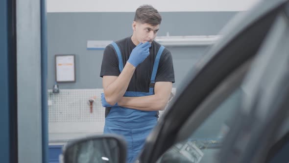 Brunette Male Auto Mechanic Touching Face and Thinking Standing in Front of Broken Vehicle in Car