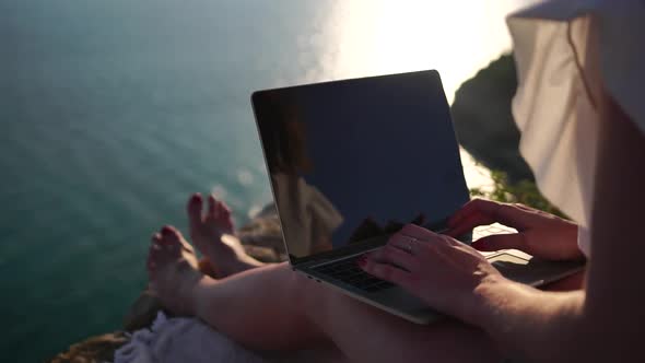 Successful Business Woman in Yellow Hat Working on Laptop By the Sea at Sunset Time