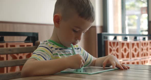 A Portrait of a Child Sits at a Table at Home and Draws on His Tablet with a Special Pen a