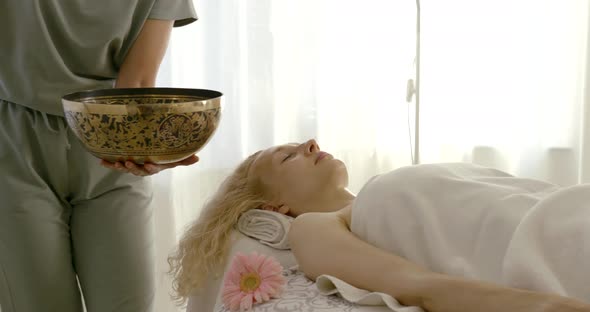 Therapist Is Holding Vibrating Tibetan Bowl Near Woman Lying on Table, Masseur Is Stroking Her Hand