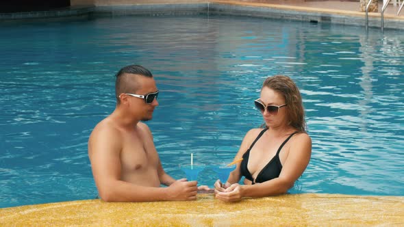 The Loving Couple Hugs and Kisses Drinking Blue Cocktail Alcohol Liquor in Swimming Pool at Hotel