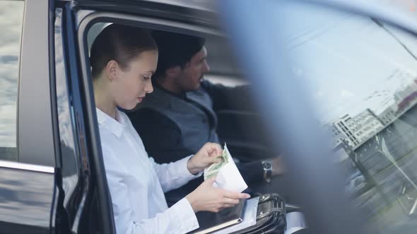 Side View Portrait of Slim Young Beautiful Woman Counting Cash Sitting in Car with Blurred Man