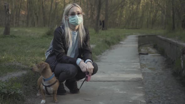 Young Blond Girl in Face Mask and Sunglasses Sitting on Hunkers in Park and Talking To Small Dog