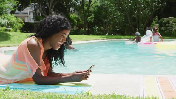 Happy african american woman relaxing at pool with family, using smartphone