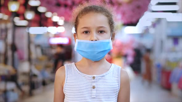 Cute Little Girl From an Epidemic of Coronaviruses or Viruses Looks at the Camera Amid Masked People