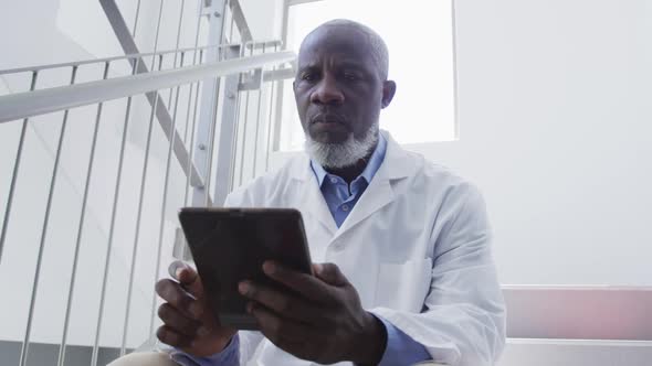 African american male doctor sitting on stairs in hospital using tablet