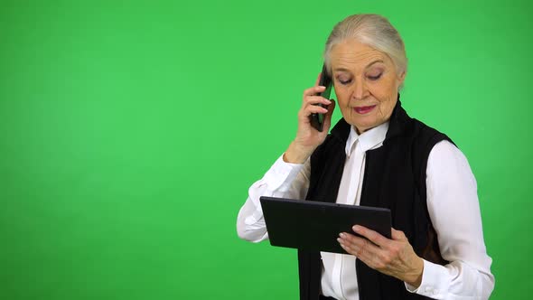 An elderly woman holds a tablet and talks on a smartphone - green screen studio