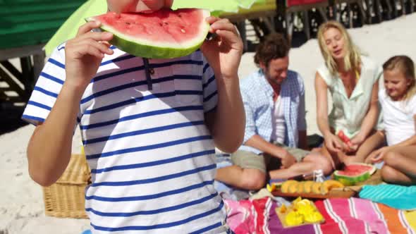 Boy eating watermelon while family sitting in background at beach