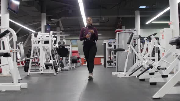 Sports Training  Africanamerican Woman Walks in the Empty Gym and Drinking Water From the Bottle