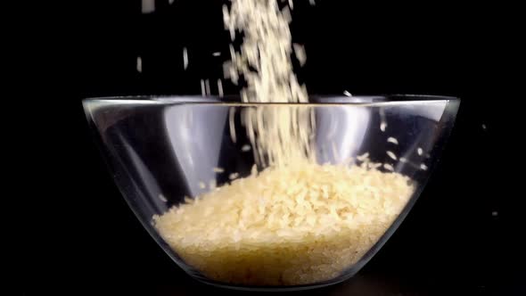 Rice is poured into a glass bowl on a black background. Slow motion.