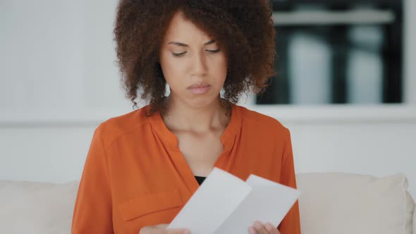 African 30s Serious Woman Sit at Couch at Home Holding Paper Reading Bad News in Letter Feels