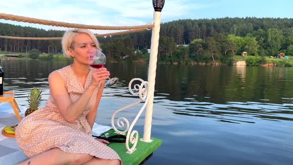 A Girl in a Sundress Sits on the Dock and Drinks Red Wine From a Glass