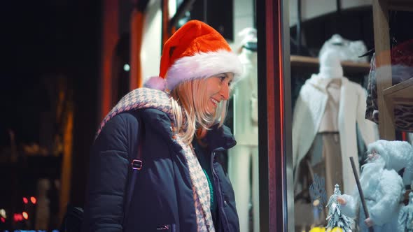 A woman in a Santa hat looks at a shop window at night.