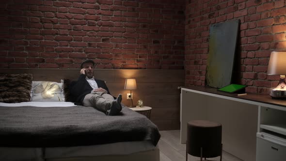 A handsome young businessman in a formal white shirt sits on a bed with a phone in a modern room.