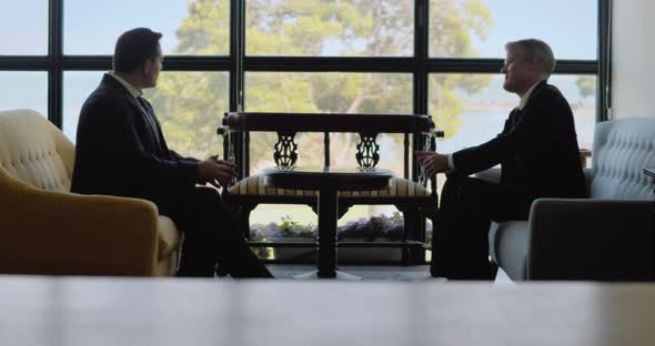 Two businessmen talking to each other while sitting in comfort sofas near a window, having a meeting