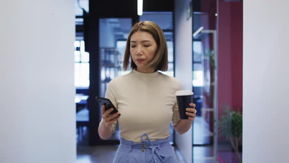 Asian businesswoman walking using smartphone holding cup of coffee in modern office